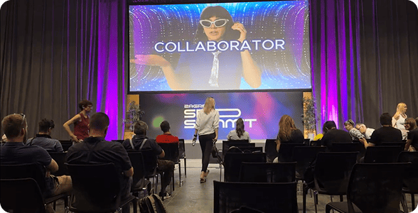 Członkowie na konferencji w Zagrzebiu, siedzący na krzesłach i zwróceni w stronę dużego ekranu wyświetlającego wideo promocyjne Collaborator.